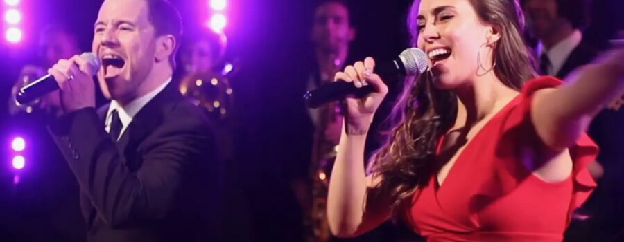 A man in a suit and a woman in a red dress sing into microphones on stage, with purple lights and musicians in the background—perfect for anyone seeking an unforgettable wedding band experience.