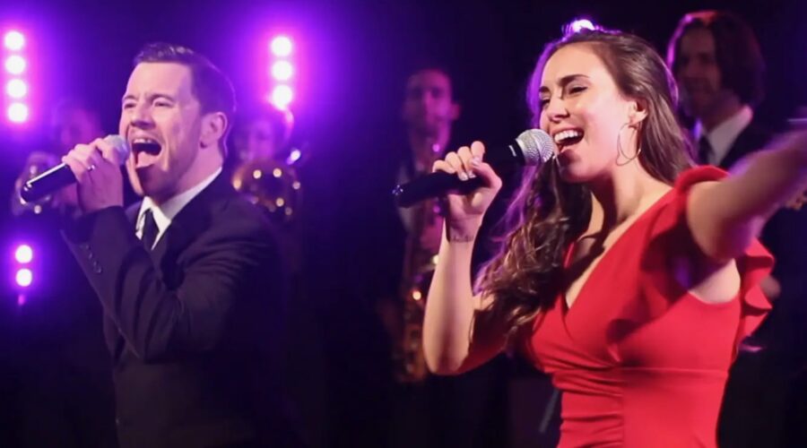 A man in a suit and a woman in a red dress sing into microphones on stage, with purple lights and musicians in the background—perfect for anyone seeking an unforgettable wedding band experience.